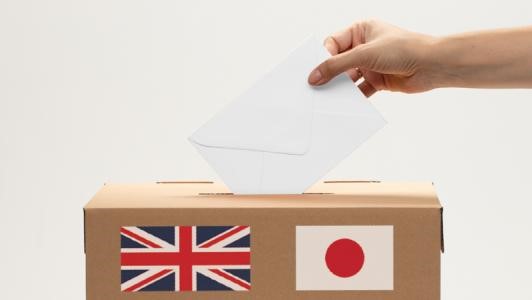 Img:Hand dropping envelope into a ballot box featuring UK and Japanese flags