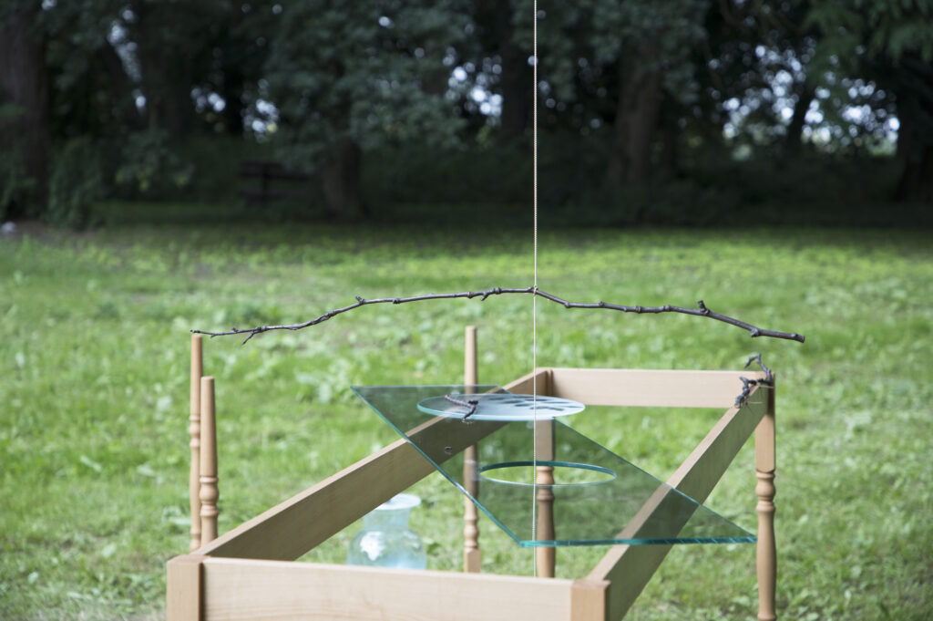 Img:Art installation of wooden frame, glass pane diagonally across it, with a string falling through a large hole in the glass