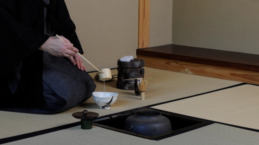 Img: Someone in kimono pouring tea into a ceramic bowl, kneeling on a tatami floor