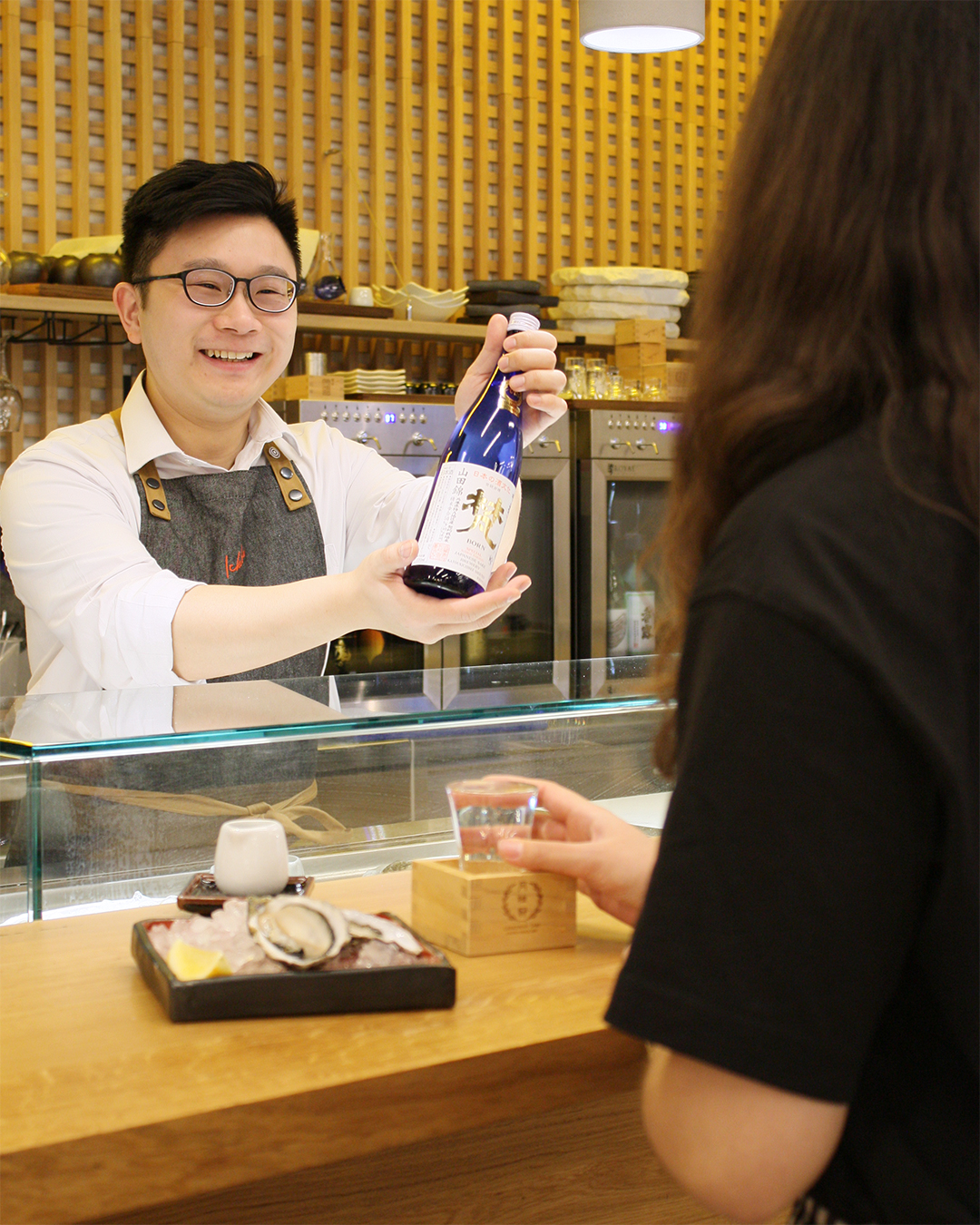 Img:man showing sake bottle at sushi counter to customer