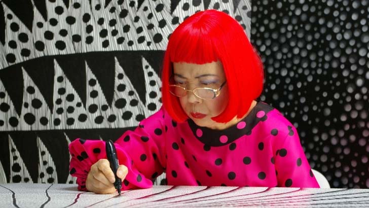 Img:Kusama working in front of her work wearing red wig and pink polcadot