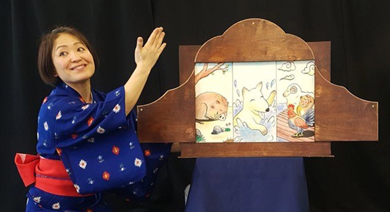 Img: Woman in kimono gesturing to a pictureboard featuring drawn animals